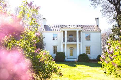 A white two-story house with a metal roof, surrounded by blooming flowers and greenery on a sunny day.