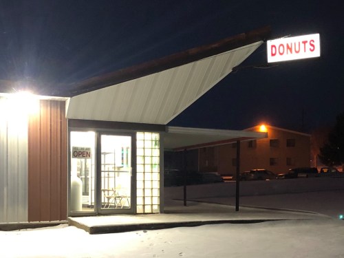 A donut shop with a bright "OPEN" sign, illuminated at night, surrounded by snow and parked cars.