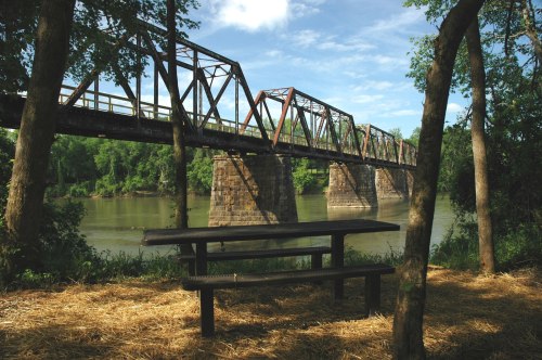 A picnic table sits on the riverbank, framed by trees, with a rusty bridge spanning the water in the background.