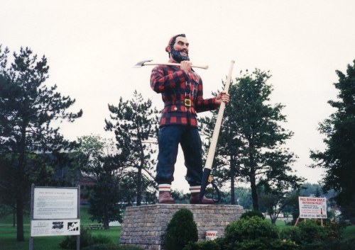 A large statue of a lumberjack in a plaid shirt, holding an axe and standing on a stone pedestal surrounded by trees.