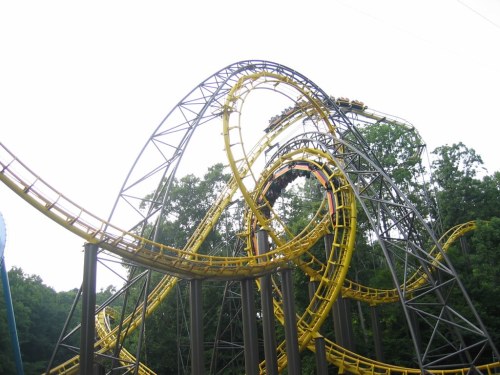 A yellow and gray roller coaster with multiple loops and twists, surrounded by green trees under a cloudy sky.
