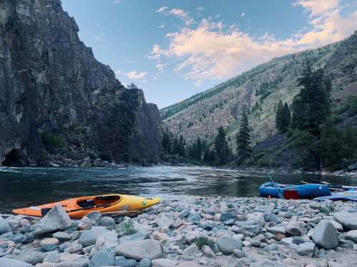 A serene river scene with rocky shores, an orange kayak, and a blue raft surrounded by mountains and trees.