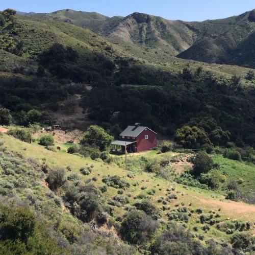 A red barn nestled in a green valley surrounded by rolling hills and mountains under a clear blue sky.