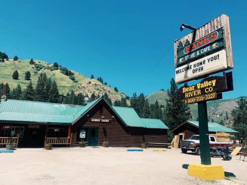 A rustic store and café with a green roof, surrounded by trees and mountains, featuring a welcoming sign.