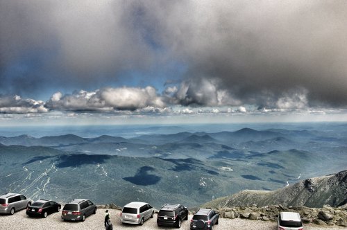 A panoramic view from a mountain summit, featuring parked cars and a dramatic sky with clouds over distant hills.