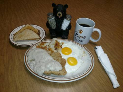 A plate with biscuits and gravy, two eggs, hash browns, toast, a coffee mug, and a bear figurine on a wooden table.
