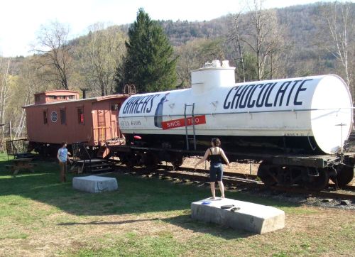 A vintage train car and a chocolate tank car on tracks, with two people nearby in a grassy area.