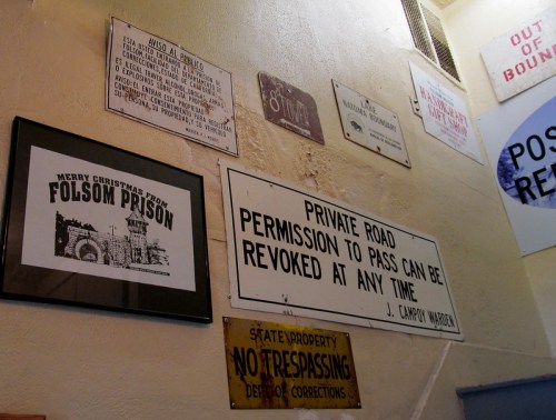 A wall displaying various signs, including a "Merry Christmas from Folsom Prison" poster and a private road warning.