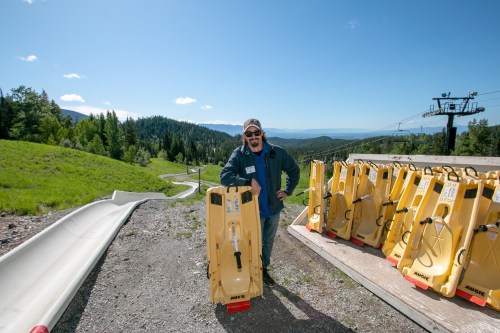 A man stands beside yellow sleds on a sunny hillside, with trees and mountains in the background.