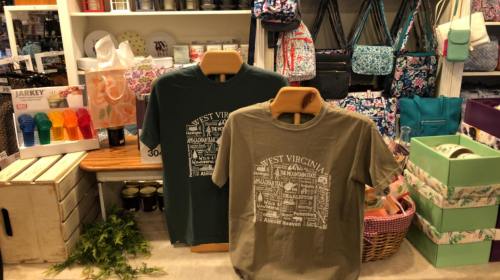 Two t-shirts displayed on wooden stands, featuring "West Virginia" designs, surrounded by various colorful accessories.