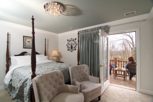 Cozy bedroom with a four-poster bed, two armchairs, and a balcony view of a deck with two people sitting outside.