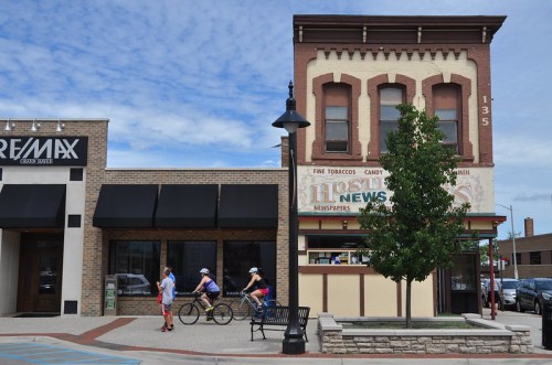 A historic building with a vintage sign, next to a modern storefront, with cyclists passing by on a sunny day.