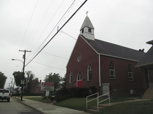 A brick church with a steeple and red door, surrounded by trees and power lines on a cloudy day.