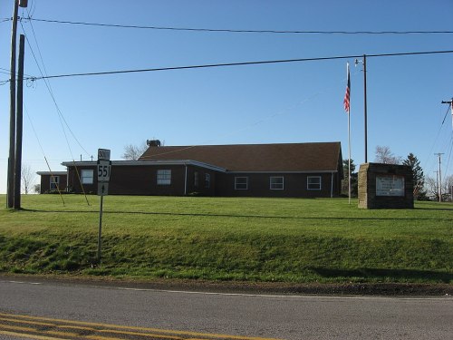 A brick building with a flagpole, set on a grassy area next to a road sign for Route 551. Clear blue sky above.