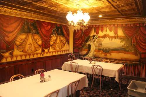 Elegant restaurant interior with ornate ceiling, decorative murals, and white tablecloths on empty tables.