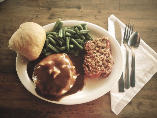 A plate of meatloaf with gravy, mashed potatoes, green beans, and a roll, accompanied by utensils on a wooden table.