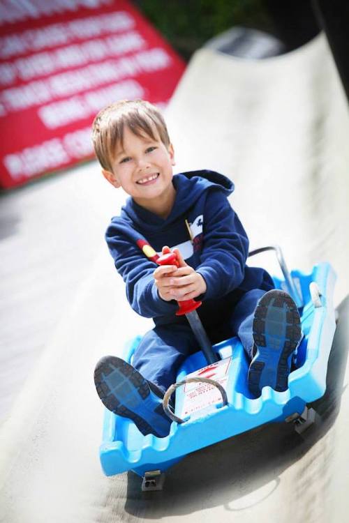 A smiling boy in a blue hoodie rides a sled down a slide, holding onto a handle with excitement.