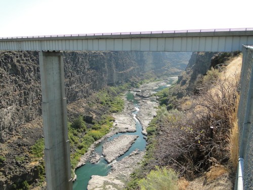 A bridge spans a deep canyon with a winding river below, surrounded by rocky cliffs and sparse vegetation.