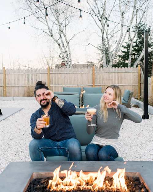 A couple enjoys drinks by a fire pit in a cozy outdoor setting, surrounded by string lights and a gravel patio.