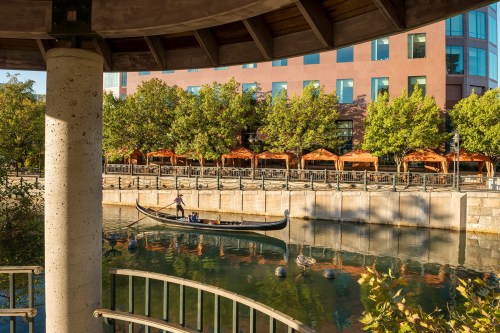 A gondola glides through a calm waterway, framed by trees and buildings, with outdoor seating visible along the shore.