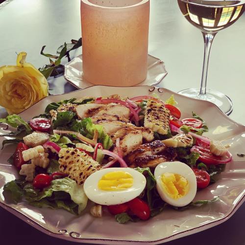 A colorful salad with grilled chicken, corn, cherry tomatoes, and boiled eggs, accompanied by a glass of white wine and a candle.