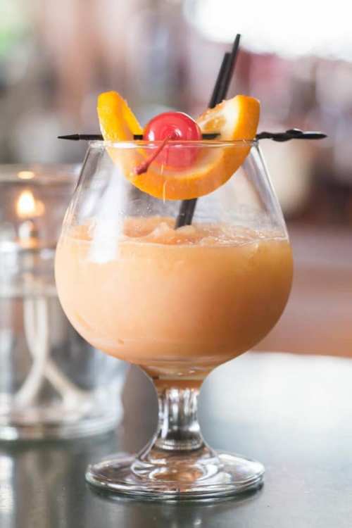 A refreshing cocktail in a glass, garnished with an orange slice and a cherry, served with a black straw.