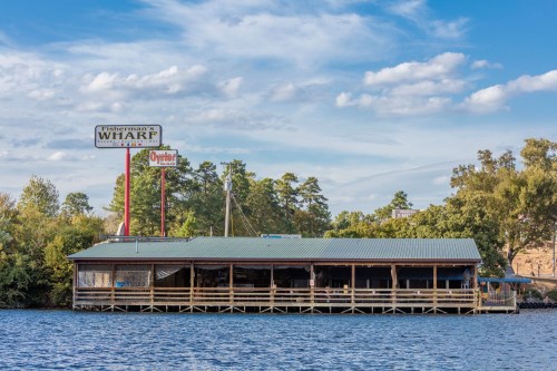 A waterfront restaurant with a green roof, wooden deck, and a sign reading "Fisherman's Wharf" against a blue sky.