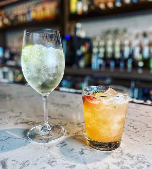 Two cocktails on a marble bar: a tall glass with ice and lime, and a short glass with ice, fruit, and a colorful drink.