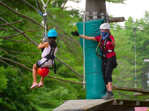 A child on a zip line approaches a platform while an instructor gestures and prepares to assist her.