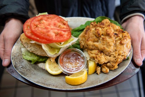A plate with a sandwich featuring lettuce, tomato, and a crab cake, served with lemon wedges and dipping sauce.
