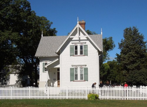 A white Victorian-style house with green shutters, surrounded by a white picket fence and trees under a clear blue sky.