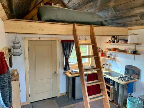 Cozy cabin interior with a lofted bed, wooden ladder, kitchenette, and rustic decor. Natural light from a window.