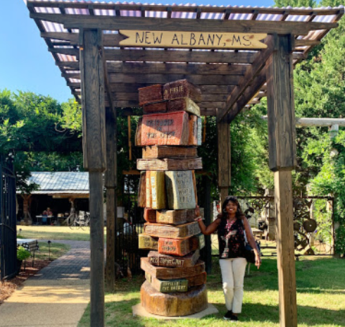 A woman stands beside a tall stack of colorful books at an entrance in New Albany, MS, under a wooden sign.