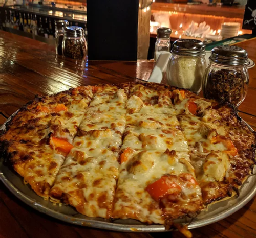 A freshly baked pizza with melted cheese, chicken, and bell peppers, served on a wooden table with condiments in the background.