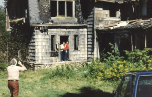 A man photographs two people standing in a doorway of an old, weathered house surrounded by overgrown grass and flowers.