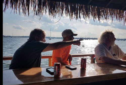 Three men sitting at a bar by the water, with one pointing towards the horizon and drinks on the table.