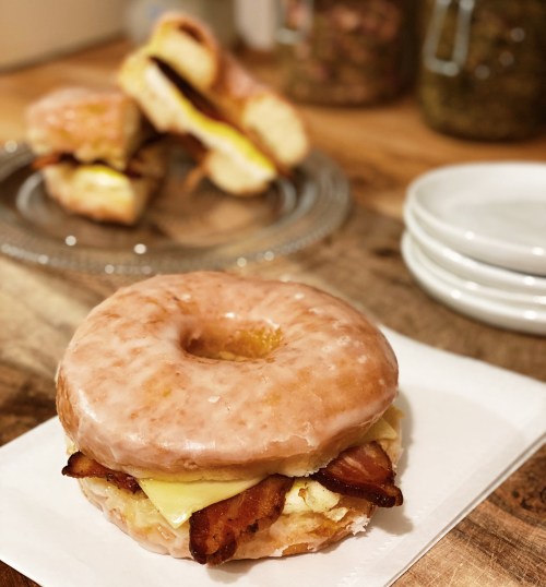 A glazed donut sandwich with bacon and cheese, served on a white plate, with more sandwiches in the background.
