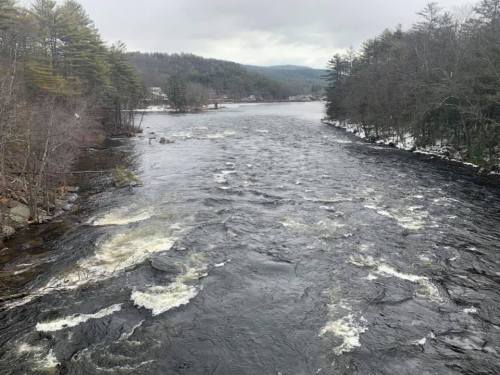 A flowing river surrounded by trees and hills under a cloudy sky, with some snow visible on the banks.