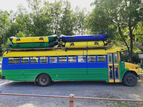 A colorful bus with green and yellow stripes, topped with two inflatable rafts, parked in a wooded area.