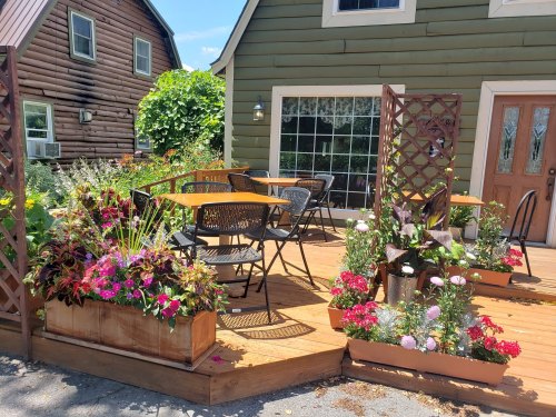 A cozy outdoor seating area with wooden tables and vibrant flower planters in front of a green house.