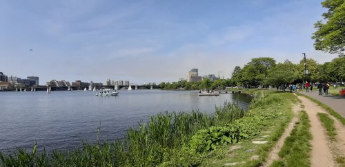 A scenic view of a river with boats, greenery, and a city skyline in the background under a clear blue sky.