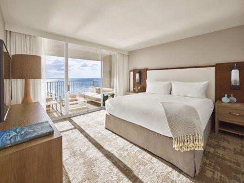 Bright hotel room with a king-sized bed, ocean view balcony, and modern furnishings. Natural light fills the space.