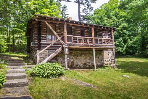 A rustic log cabin with a stone foundation, surrounded by lush greenery and trees, featuring a porch with wooden railings.