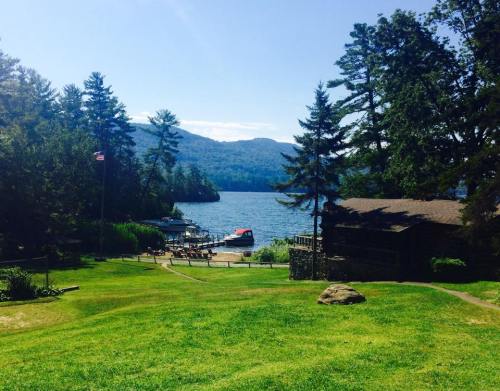 A serene lakeside view with boats, trees, and a clear blue sky, framed by green grass and a rustic cabin.