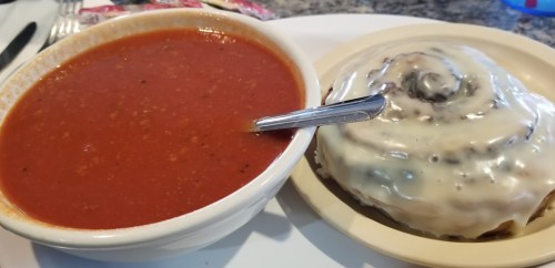 A bowl of rich tomato soup next to a large cinnamon roll topped with cream cheese icing.