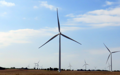 A tall wind turbine stands in a field, surrounded by more turbines under a blue sky with scattered clouds.