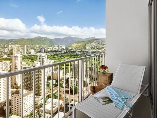 A balcony with a lounge chair overlooks a cityscape and mountains under a blue sky with clouds.