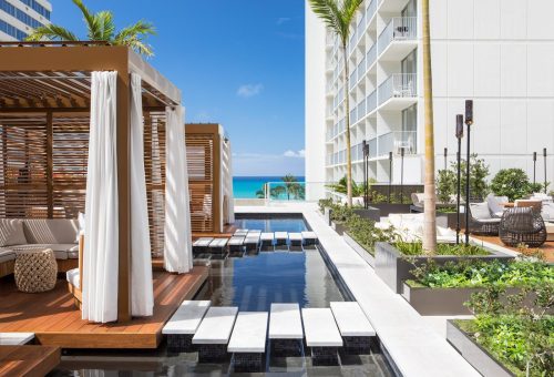 A modern outdoor lounge area with cabanas, a pool, and ocean views, surrounded by tropical plants and bright blue skies.