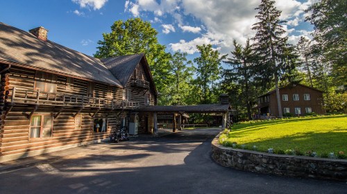 A rustic log cabin surrounded by trees, with a grassy area and a stone wall along the driveway.
