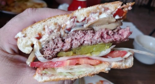 A close-up of a hand holding a juicy hamburger with layers of lettuce, tomato, pickles, and mayonnaise on toasted bread.
